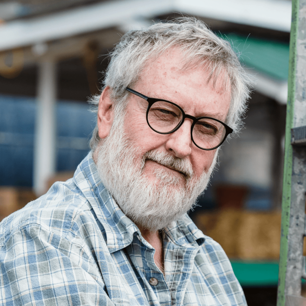 Ein älterer Herr mit vollem grauen Bart und grauen Haaren, der eine runde Brille trägt. Er lächelt freundlich und trägt ein kariertes Hemd in Blautönen. Der Hintergrund ist unscharf, aber es ist ein Teil eines Gebäudes zu erkennen.